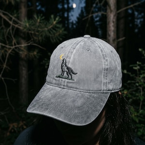 May include: Gray baseball cap featuring an embroidered wolf howling at a yellow crescent moon. The cap is made of a washed, textured fabric. The background shows a forest scene with trees and a moon.
