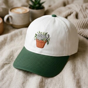 May include: White baseball cap with a green brim and button. The cap features an embroidered design of a gray cat in a terracotta pot with green leaves. The cap is a stylish accessory.