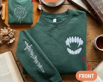 May include: A dark green sweatshirt with white embroidered designs. The front features a stylized fang design, the sleeve has a sword, and the back has a circular design. The sweatshirt is displayed on a wooden surface with books and teacups.