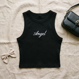 May include: A black ribbed tank top featuring the word Angel embroidered in white cursive script across the chest. The garment has a round neckline and sleeveless cut. A small beige hair bow and a black shoulder bag are positioned nearby.