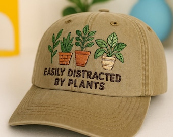 May include: A khaki baseball cap with an embroidered design of three potted plants and the text "EASILY DISTRACTED BY PLANTS" in brown thread. The cap has a curved brim and a classic six-panel design.