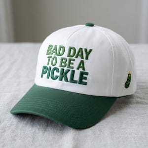 May include: White baseball cap with a green brim and top button. The cap features the embroidered phrase "BAD DAY TO BE A PICKLE" in green, along with a small embroidered pickle on the side. A fun, casual accessory.