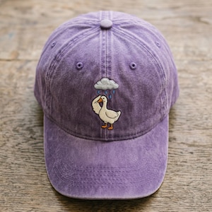 May include: A purple baseball cap with a curved brim. The cap features an embroidered design of a white goose under a cloud with raindrops. The goose is raising its wing. The cap is made of a soft, textured material.