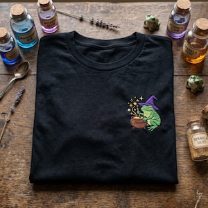 May include: Black t-shirt with an embroidered design of a green frog wearing a purple witch hat, stirring a cauldron. The design includes gold stars and a bubbling potion. The shirt is surrounded by potion bottles and decorative items.