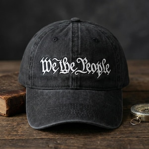May include: A charcoal black baseball cap with a washed, vintage-style finish. The front panel features the text "We the People" embroidered in white thread using a decorative, gothic-inspired script font. The cap has a curved brim and eyelets.