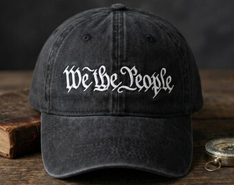 May include: A charcoal black baseball cap with a washed, vintage-style finish. The front panel features the text "We the People" embroidered in white thread using a decorative, gothic-inspired script font. The cap has a curved brim and eyelets.