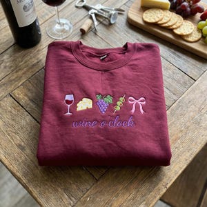 May include: A maroon sweatshirt with embroidered wine-themed graphics. The design includes a wine glass, cheese, grapes, olives, and a bow, with the words "wine o' clock" in cursive. The sweatshirt is folded on a wooden surface.