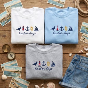 Könnte beinhalten: Drei Sweatshirts mit Rundhalsausschnitt in Weiß, Hellblau und Grau, jeweils mit einem nautischen Design und den Worten "harbor days" bestickt. Das Design umfasst eine Möwe, einen Leuchtturm, einen Anker, ein Seil und ein Segelboot. Postkarten und ein Kompass sind im Hintergrund.