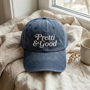 May include: A navy blue baseball cap featuring the embroidered words "Pretti & Good" in white script. The cap is displayed on a beige fabric, with a neutral-toned mug and dried flowers in the background.