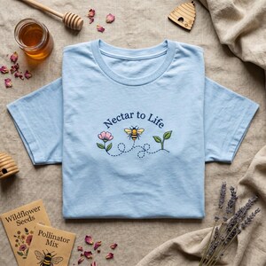 May include: A light blue t-shirt featuring an embroidered design of a pink flower, a yellow and black bee, and a green sprout. The text "Nectar to Life" is arched above the bee, with a dotted line tracing the bee's flight path across the chest.