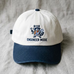 May include: A white and navy blue baseball cap featuring a cartoon raccoon holding a wrench and blueprints. The text "ENGINEER MODE" is below the raccoon graphic. The cap has a curved peak and a button on top.