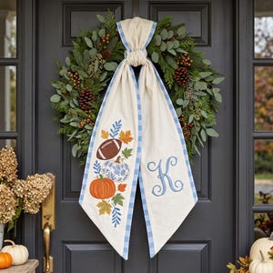 May include: A decorative wreath featuring green foliage, pinecones, and berries, adorned with a long, cream-colored fabric sash. The sash has a blue gingham border and displays embroidered autumn leaves, a football, a pumpkin, and the letter K.