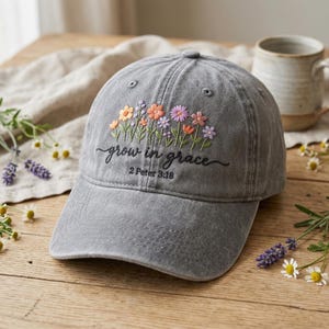 May include: A charcoal gray baseball cap featuring embroidered colorful wildflowers in shades of orange, pink, and purple. The text "grow in grace" is stitched in black cursive script above the text "2 Peter 3:18" in a simple font.
