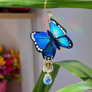 Può includere: Un orecchino pendente a forma di farfalla blu con accenti di cristallo. La farfalla è realizzata in resina e ha un contorno nero. L'orecchino è appeso a un cerchio dorato.