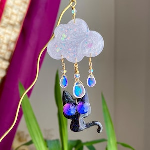 May include: A whimsical handmade bookmark featuring a black cat with iridescent eyes and a cloud. The cloud is white with glitter, and the raindrops are iridescent. The bookmark is suspended from a gold wire.