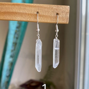 May include: A pair of silver earrings with clear quartz crystal drops. The earrings are hanging from a wooden display stand. The number "1" is written on the stand.