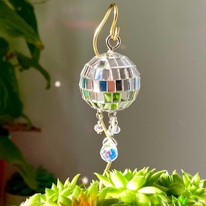 Może przedstawiać: Mały, srebrny ornament w kształcie kuli dyskotekowej z wiszącymi kryształowymi kroplami. Ornament jest przymocowany do złotego drutu i znajduje się w doniczce z zielonymi sukulentami.