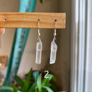 May include: A pair of silver earrings with clear quartz crystal pendants. The earrings are hanging from a wooden display stand.