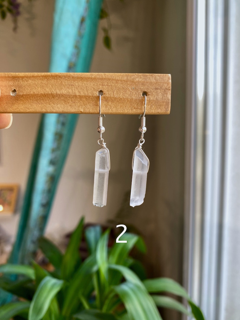 May include: A pair of silver earrings with clear quartz crystal pendants. The earrings are hanging from a wooden display stand.