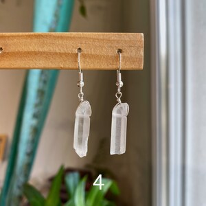 May include: A pair of silver earrings with clear quartz crystal pendants. The crystals are long and slender, with a slightly pointed tip. The earrings are hanging from a wooden display stand.