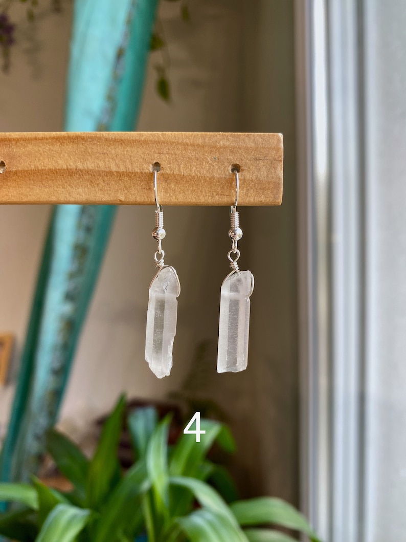 May include: A pair of silver earrings with clear quartz crystal pendants. The crystals are long and slender, with a slightly pointed tip. The earrings are hanging from a wooden display stand.