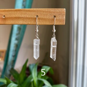 May include: A pair of silver earrings with clear quartz crystal pendants. The crystals are faceted and have a natural, raw look. The earrings are hanging from a wooden display stand.