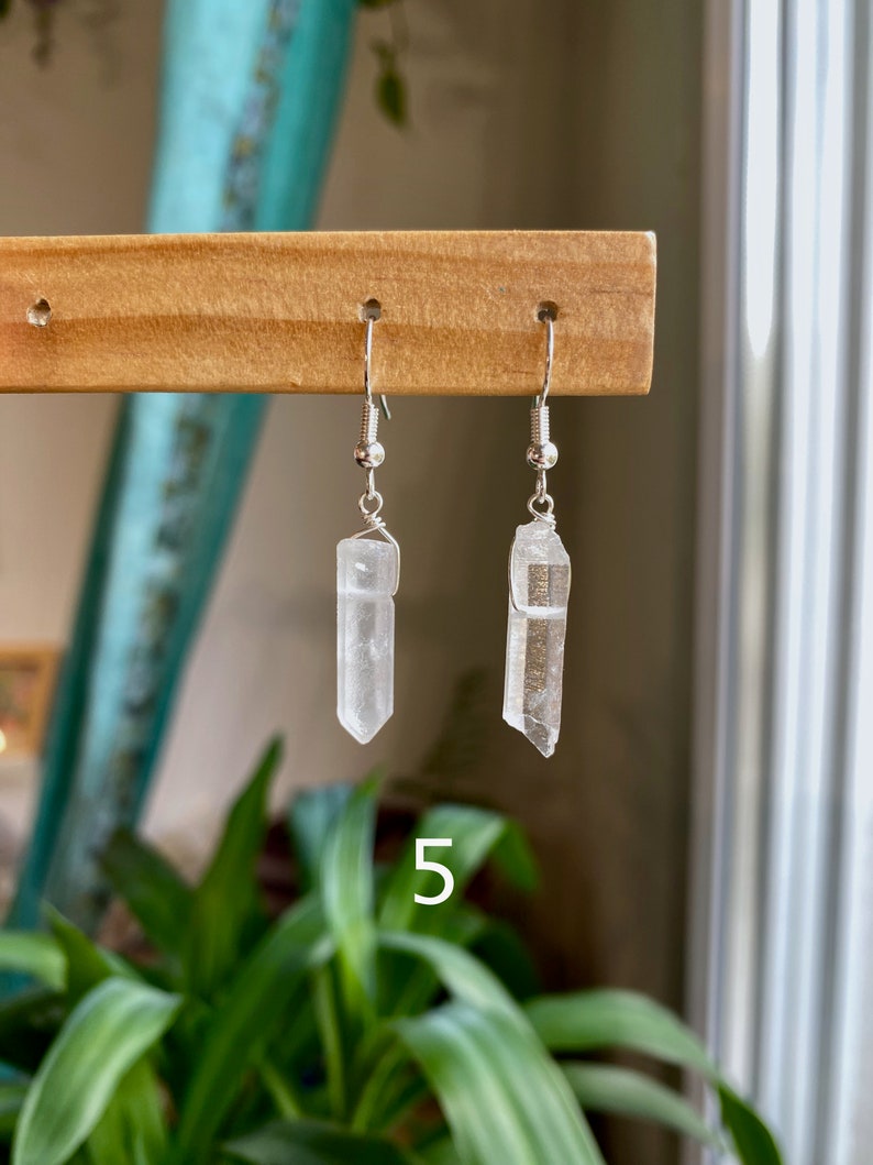 May include: A pair of silver earrings with clear quartz crystal pendants. The crystals are faceted and have a natural, raw look. The earrings are hanging from a wooden display stand.