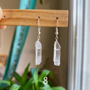 May include: A pair of silver earrings with clear quartz crystal pendants. The crystals are long and slender, with a slightly pointed tip. The earrings are hanging from a wooden display stand.