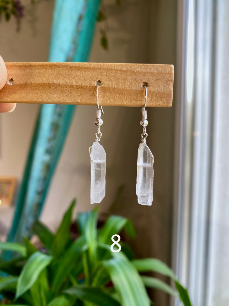 May include: A pair of silver earrings with clear quartz crystal pendants. The crystals are long and slender, with a slightly pointed tip. The earrings are hanging from a wooden display stand.
