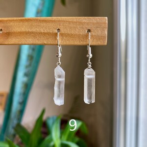 May include: A pair of silver earrings with clear quartz crystal pendants. The crystals are long and slender, and the earrings are hanging from a wooden display stand.