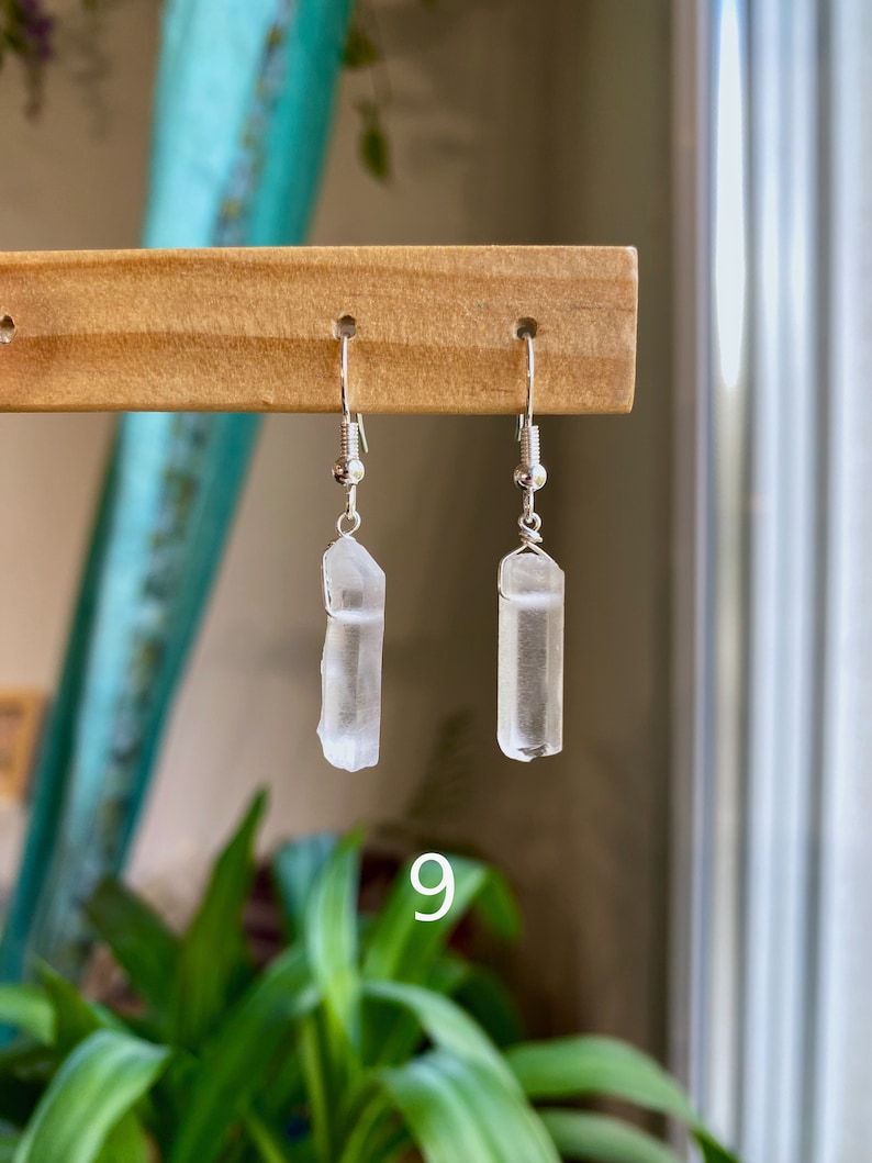 May include: A pair of silver earrings with clear quartz crystal pendants. The crystals are long and slender, and the earrings are hanging from a wooden display stand.