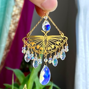 Puede incluir: Un adorno de metal dorado con forma de polilla que presenta intrincados patrones recortados y fases lunares. La pieza está decorada con múltiples cristales transparentes e iridiscentes en forma de lágrima y esfera que cuelgan de los bordes.