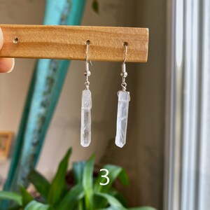 May include: A pair of silver earrings with clear crystal point pendants. The earrings are hanging from a wooden display stand.