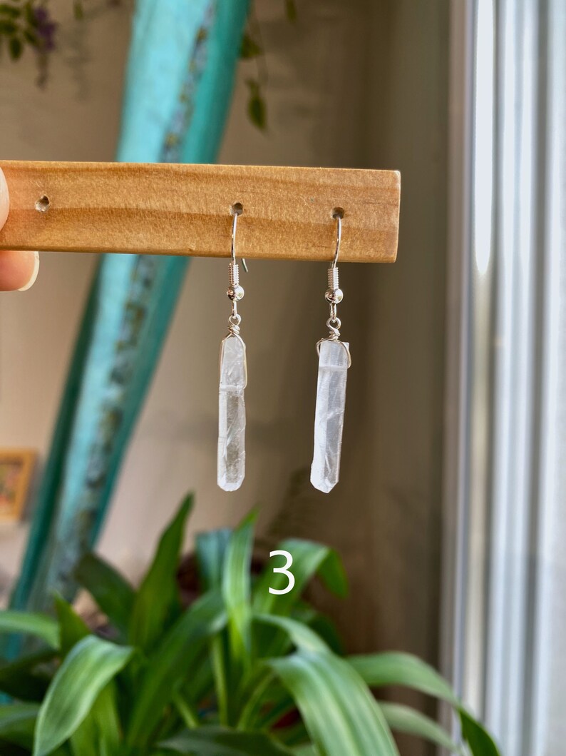 May include: A pair of silver earrings with clear crystal point pendants. The earrings are hanging from a wooden display stand.
