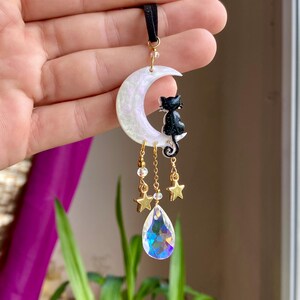 May include: A celestial-themed pendant featuring a pearlescent crescent moon with a black cat silhouette. Gold-colored stars and a teardrop-shaped crystal dangle below. The pendant is suspended from a black cord.