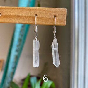May include: A pair of silver earrings with clear quartz crystal pendants. The crystals are long and slender, with a slightly pointed tip. The earrings are hanging from a wooden display stand.
