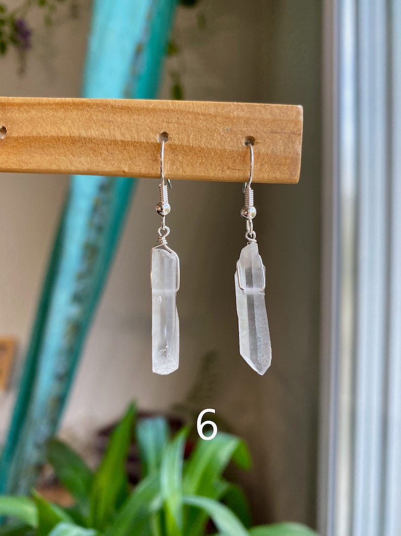 May include: A pair of silver earrings with clear quartz crystal pendants. The crystals are long and slender, with a slightly pointed tip. The earrings are hanging from a wooden display stand.