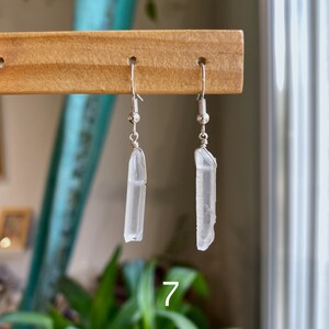 May include: A pair of silver earrings with clear quartz crystal pendants. The crystals are long and slender, with a natural, raw look. The earrings are hanging from a wooden display stand.