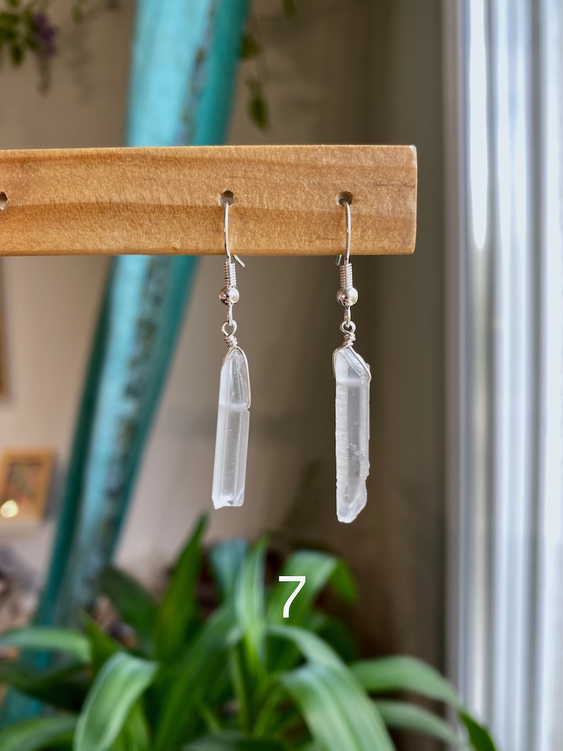 May include: A pair of silver earrings with clear quartz crystal pendants. The crystals are long and slender, with a natural, raw look. The earrings are hanging from a wooden display stand.