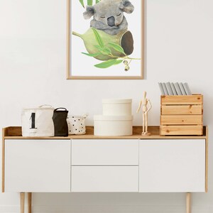 May include: A framed watercolor print of a sleeping koala bear nestled in a eucalyptus branch. The artwork features soft gray and green tones, creating a calming aesthetic. The print is displayed above a white and wood-toned cabinet.