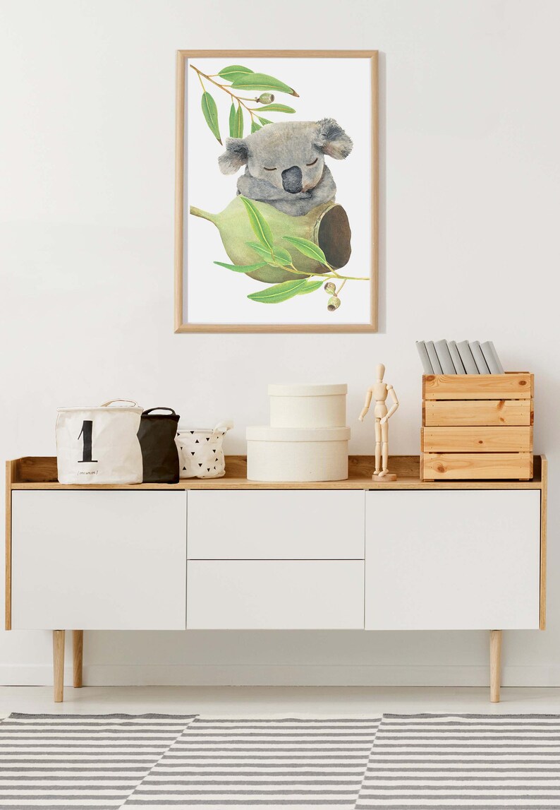 May include: A framed watercolor print of a sleeping koala bear nestled in a eucalyptus branch. The artwork features soft gray and green tones, creating a calming aesthetic. The print is displayed above a white and wood-toned cabinet.
