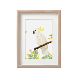 May include: A framed watercolor print featuring a white cockatoo perched on a brown branch. The bird has yellow crest feathers and a black beak. Green leaves and yellow flowers surround the bird. The frame is light brown.