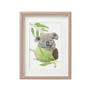 May include: Watercolor print of a sleeping koala bear nestled in a eucalyptus branch. The artwork features soft gray and green tones, framed in a light wood frame with a white mat. The koala is depicted with its eyes closed, resting on a green pod.