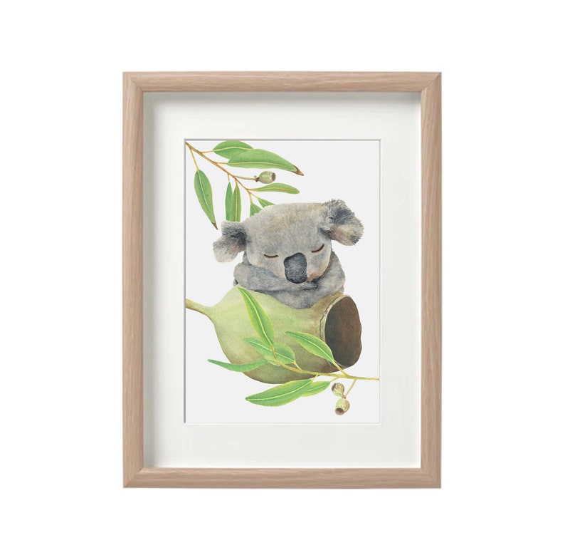 May include: Watercolor print of a sleeping koala bear nestled in a eucalyptus branch. The artwork features soft gray and green tones, framed in a light wood frame with a white mat. The koala is depicted with its eyes closed, resting on a green pod.