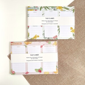 May include: Two task planners with watercolour floral and fruit designs. The top planner has a green and yellow floral pattern, while the bottom one features strawberries and oranges. Each planner has sections for goals, to-do lists, and self-care, with the text "YOU'VE GOT THIS!" at the top.