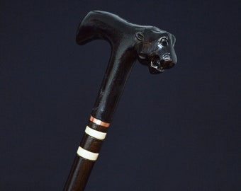 May include: A dark brown wooden walking stick with a carved animal head at the top. The stick has white and copper accents.