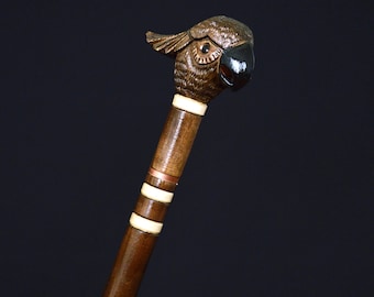 May include: A wooden walking stick with a carved parrot head handle. The handle is brown with black eyes and a black beak. The stick is brown with white and copper accents.