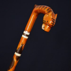 Op de afbeelding: Een houten wandelstok met een gegraveerd tijgerhoofd aan de bovenkant. De stok is geschilderd met bruine, oranje en witte strepen.