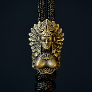 Op de afbeelding: Een gouden hanger met een gedetailleerd buste van een vrouw met een hoofdtooi. De hanger is bevestigd aan een zwart en gouden koord.