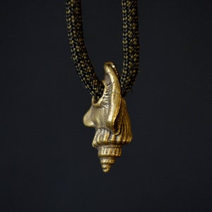 Puede incluir: Un colgante de concha de mar de color dorado que cuelga de un cordón negro. El colgante es detallado y tiene una forma espiral.